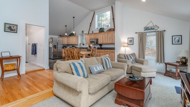 A spacious living area with a large sofa, a coffee table, and a dining table. There is a kitchen in the background with wooden cabinets and a refrigerator.