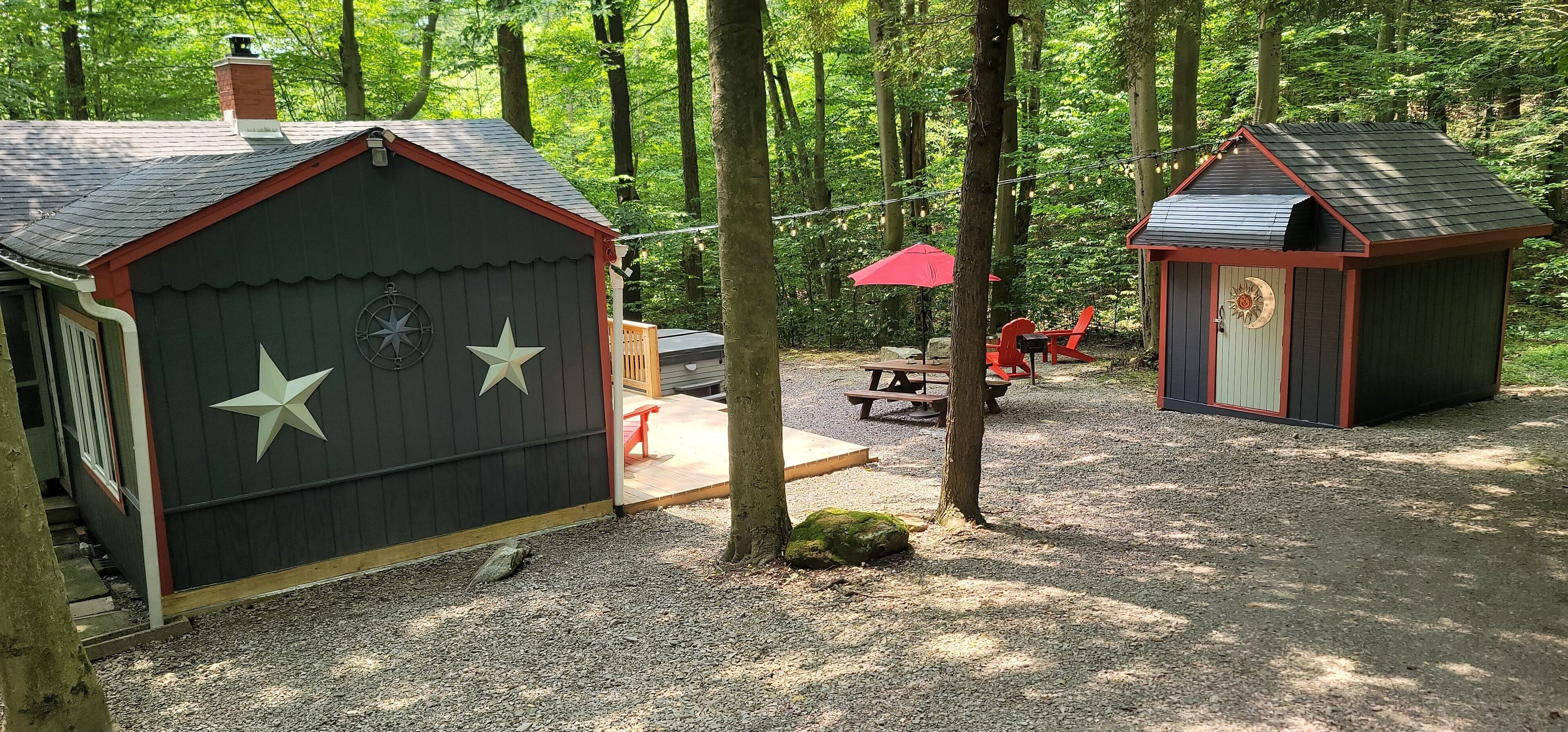 A forested campsite with two small cabins, picnic tables, and a red umbrella.