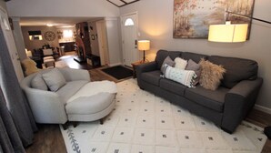 Ein Wohnzimmer mit grauem Sofa, grauem Sessel und weißem Teppich. An der Wand hängt ein Bild und auf einem Beistelltisch steht eine Lampe.