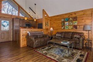 A cozy cabin interior with wooden walls, a sofa set, a coffee table, and a kitchen area.