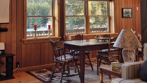 A cozy cabin interior with a large dining table, wooden walls, and a view of trees through large windows.