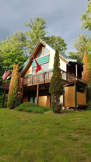 A two-story wooden building with a balcony, surrounded by greenery and trees. - Loon Lake, Chestertown - spotless, bright chalet for  6-8, close to Public Beach (Chestertown)