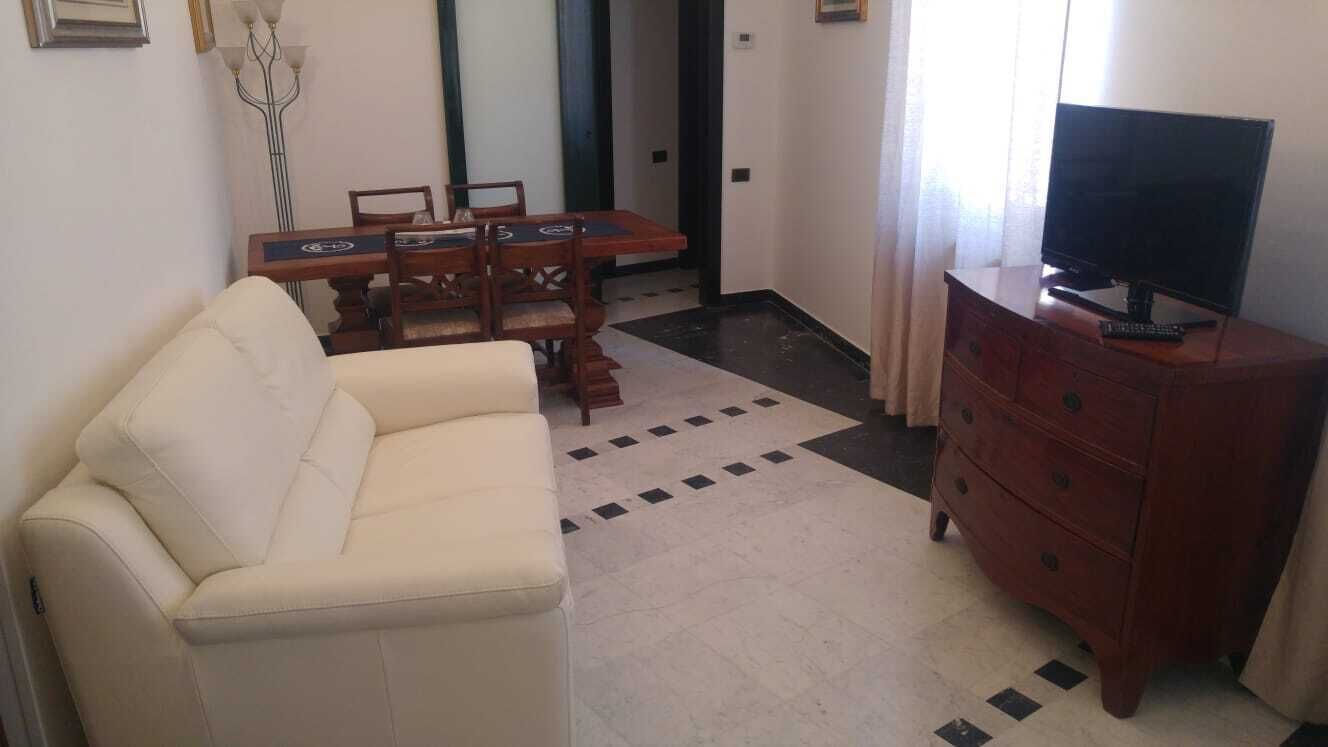 A living room with a beige sofa, a wooden dining table with chairs, a TV on a wooden cabinet, and a patterned rug.