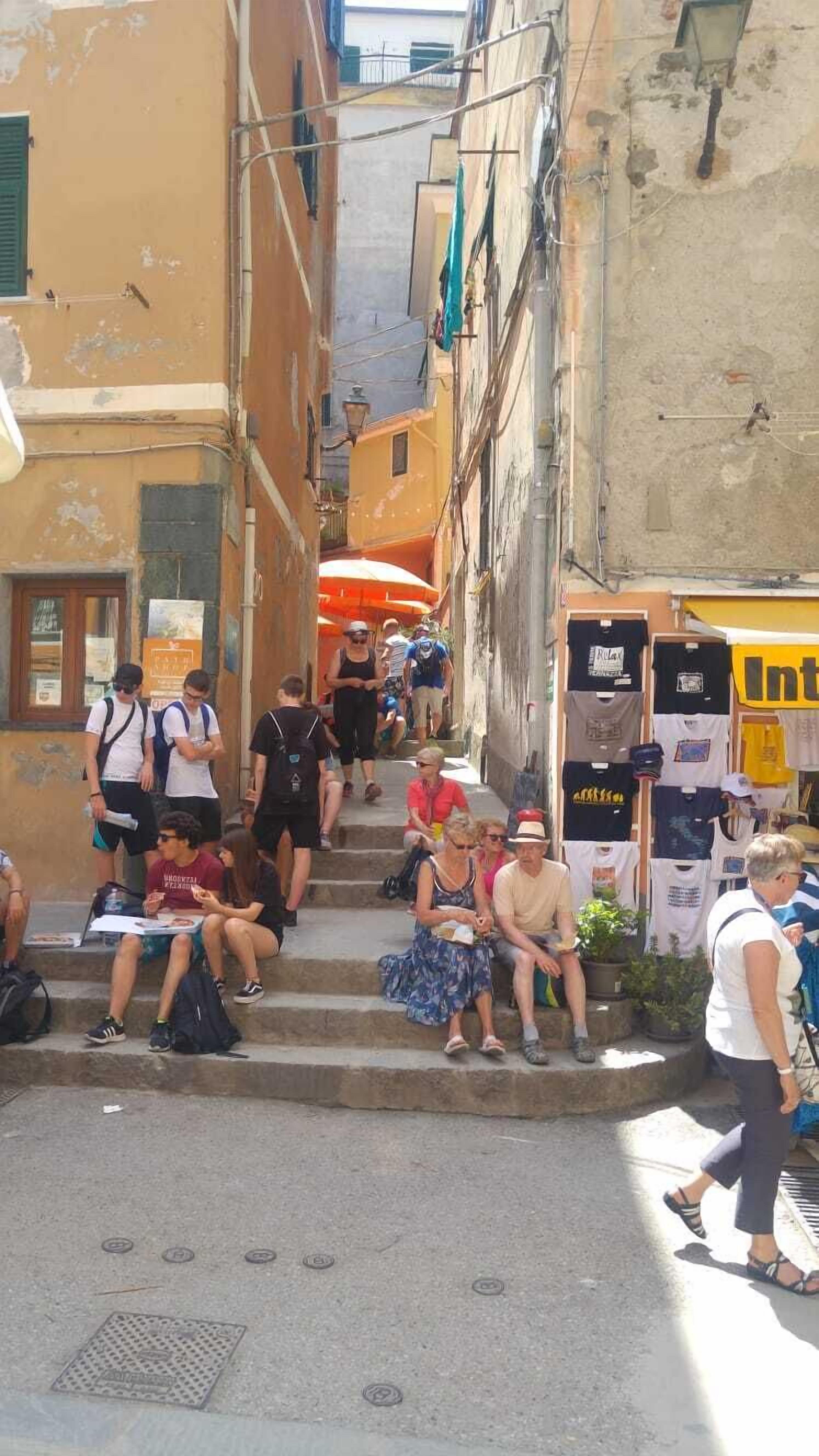 A narrow street with steps leading up to a building. People are sitting and walking on the steps.