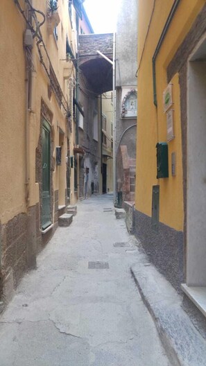 Une ruelle étroite, bordée de bâtiments des deux côtés, dont les façades présentent un mélange de pierres et de crépi, et qui, pour certaines, montrent des signes d’usure.