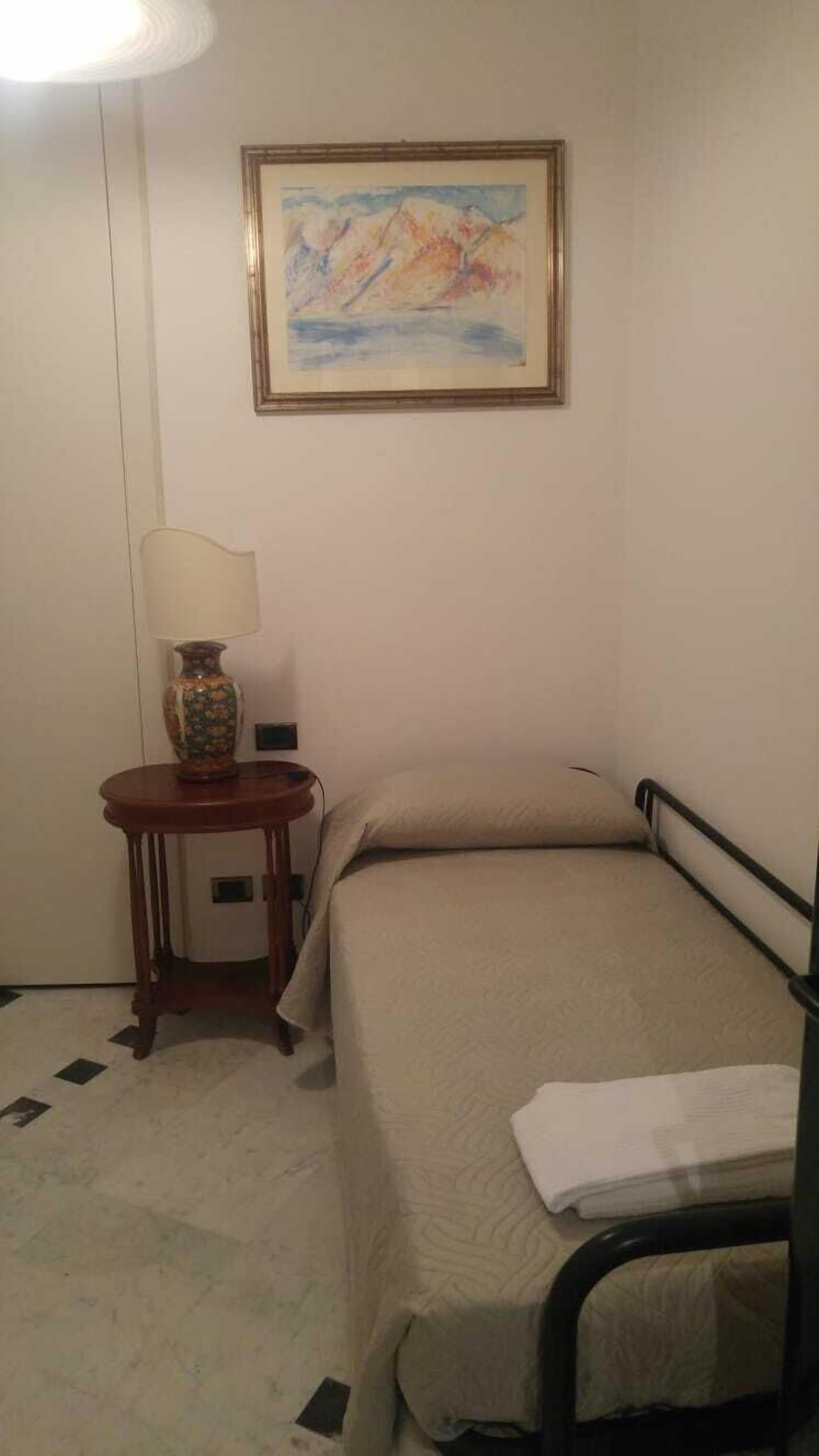 A small, neatly arranged bedroom with a single bed, a bedside table with a lamp, and a framed painting on the wall.