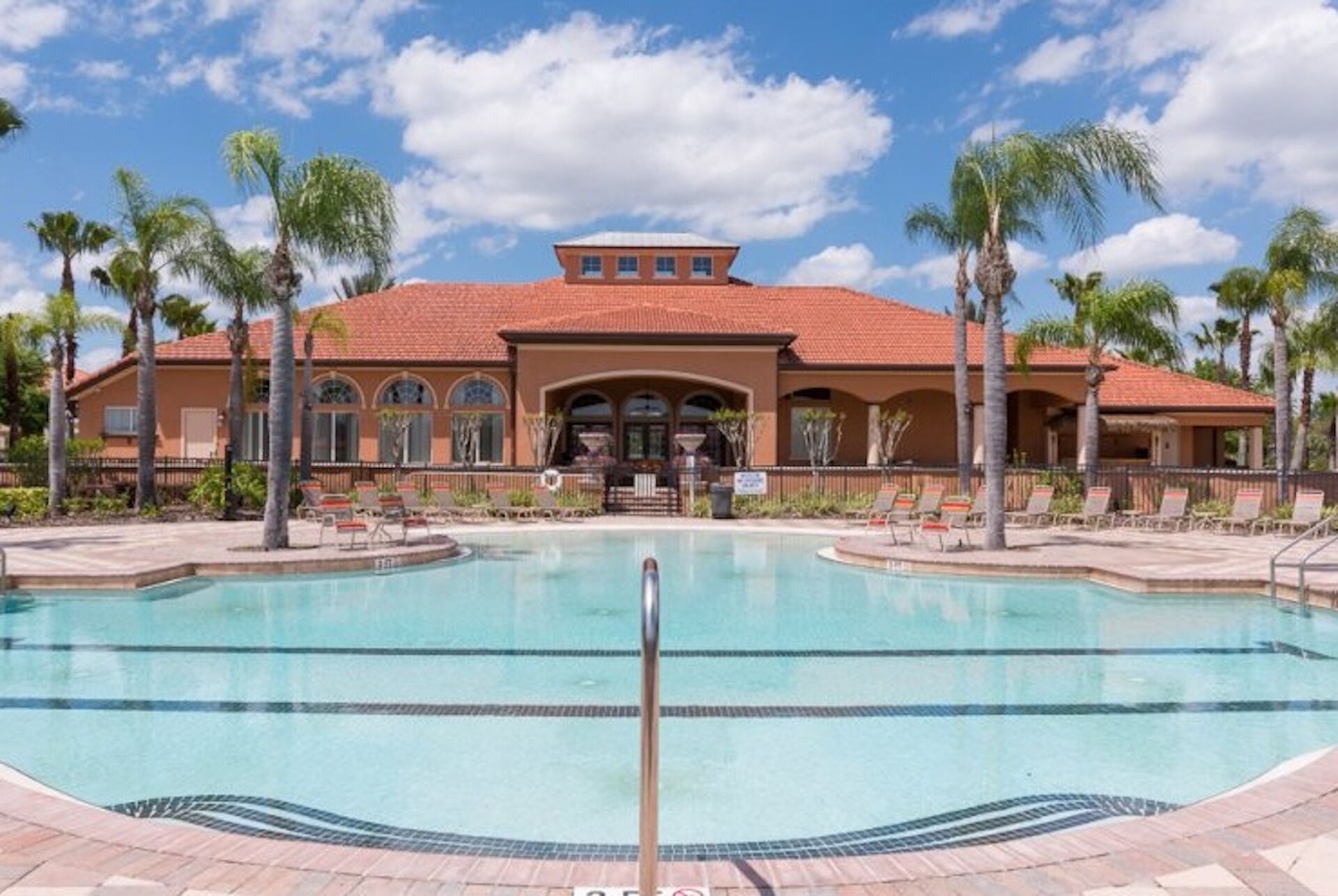 Een resort met een groot zwembad, palmbomen en een gebouw met een rode dakbedekking.