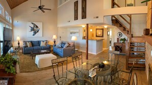 A spacious living area with a high ceiling, a ceiling fan, a large sofa set, a glass dining table, and a staircase leading to an upper level.