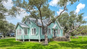 Ein zweistöckiges Haus mit grüner Fassade, einer Holzveranda und einem großen Baum davor.