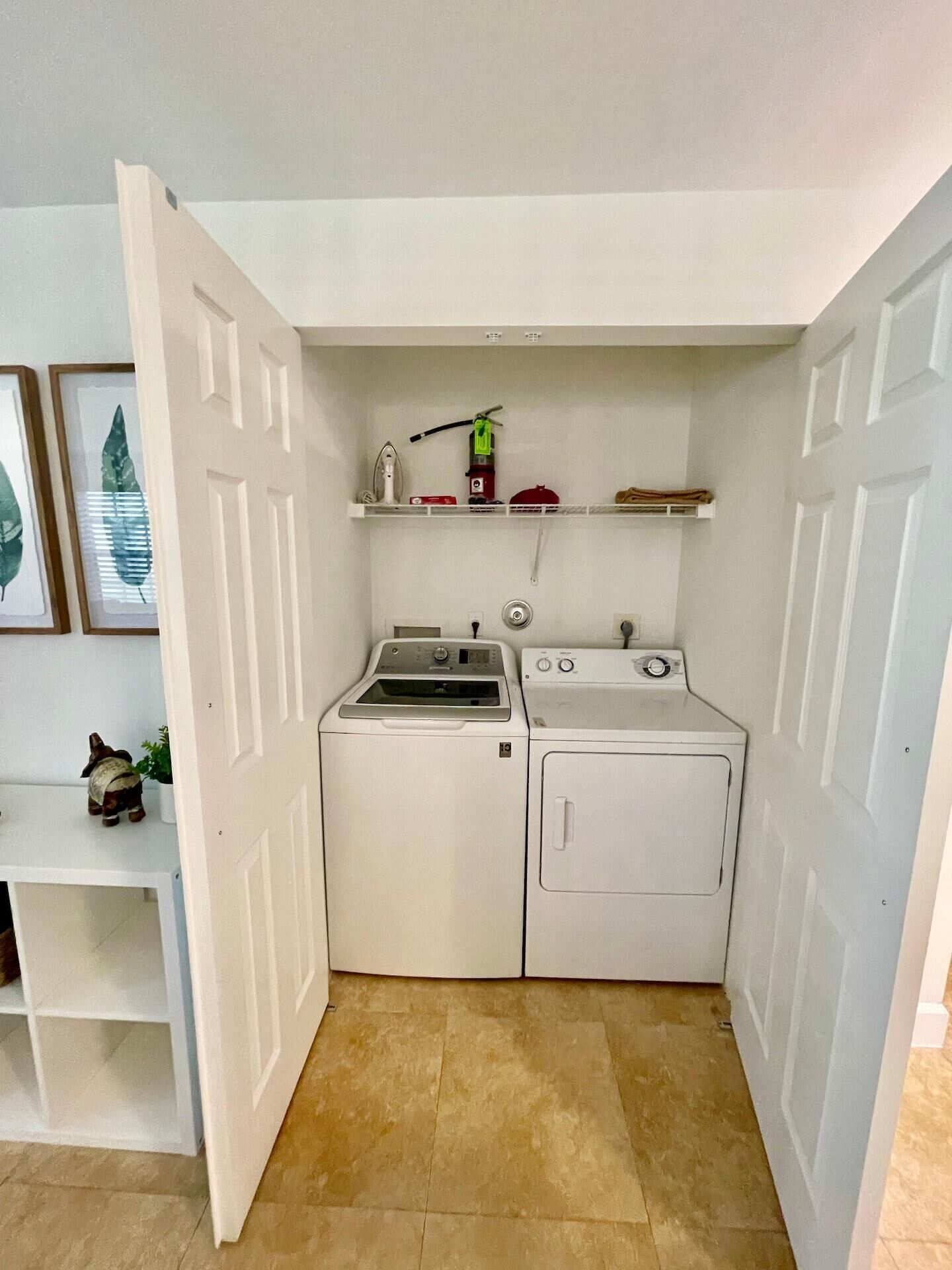 A laundry room with two washing machines and a dryer, a shelf with cleaning supplies, and a small shelf with decorative items.