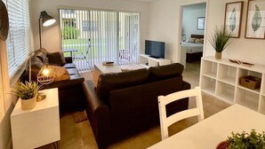 A modern living room with a sofa, TV, and dining area. There is a large window with blinds, a ceiling light fixture, and a plant on a shelf. - 360A - Boca Retreat w/ Pool, Parking, Near Evert Tennis & Shops (Boca Raton)