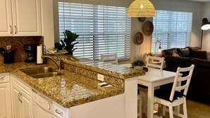 A kitchen with a granite countertop, white cabinets, a dining area with a table and chairs, and a large window with blinds. - 360A - Boca Retreat w/ Pool, Parking, Near Evert Tennis & Shops (Boca Raton)