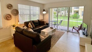 A modern living room with a sectional sofa, a coffee table, and a TV. There are large windows with blinds, a floor lamp, and a view of a garden outside. - 360A - Boca Retreat w/ Pool, Parking, Near Evert Tennis & Shops (Boca Raton)