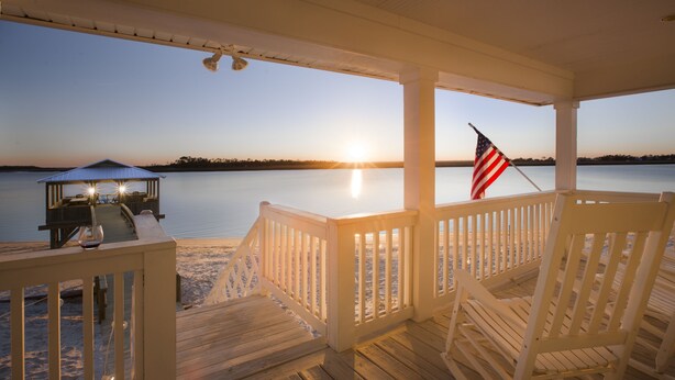 A wooden deck with a view of the sunset over water, a covered seating area, and an American flag. - Beach Front South End, Designer Decorated, Dream Retreat on Tybee Island! (Tybee Island)