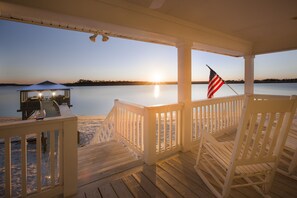 Une terrasse en bois offrant une vue sur le coucher de soleil sur l’eau, un espace salon abrité et un drapeau américain.