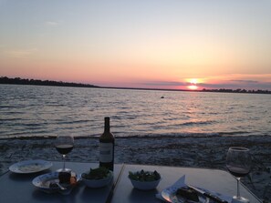 Une table dressée pour un repas, avec une bouteille de vin, des verres et des assiettes remplies de nourriture, au bord d’un lac, au coucher du soleil.