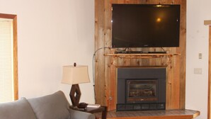 A cozy living room with a fireplace, a flat-screen TV mounted above it, a grey sofa, and a small table with a lamp.
