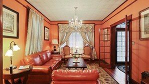 Une pièce avec un canapé en cuir rouge, une table basse en bois et un lustre. On y trouve deux fauteuils, une lampe et une fenêtre avec des rideaux.