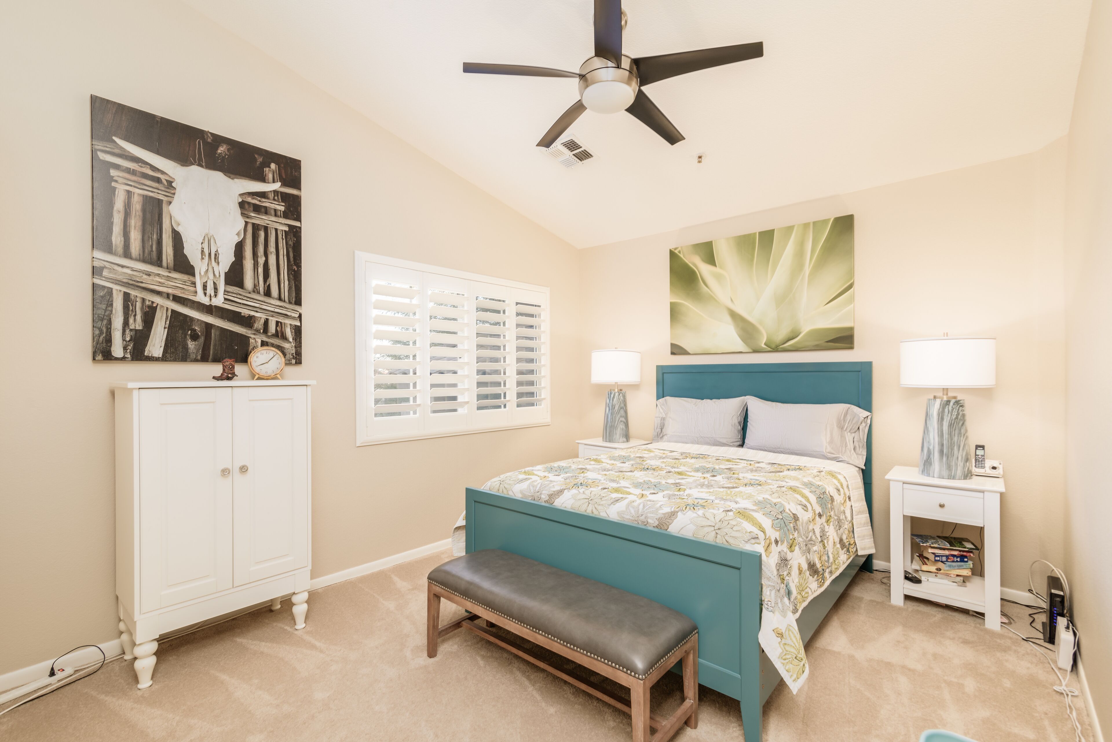 Une chambre à coucher avec un lit de couleur turquoise, des tables de chevet blanches, un ventilateur de plafond, une armoire blanche et une grande fenêtre avec des stores.