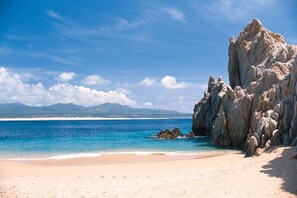 Uma praia com águas turquesa cristalinas, um afloramento rochoso e montanhas ao longe.
