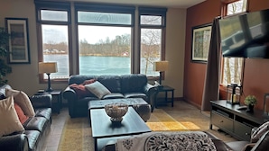 Un salon avec un canapé en cuir, une table basse et un téléviseur posé sur un meuble. De grandes fenêtres offrent une vue sur un lac et des arbres.