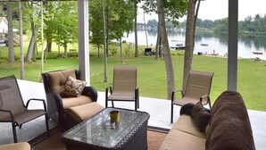 Une terrasse couverte, aménagée avec des meubles en osier, une table en verre et offrant une vue sur un lac.