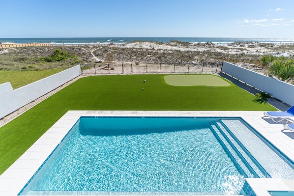 Ein Pool mit klarem, blauem Wasser, eine grüne Rasenfläche und Blick auf Strand und Meer.