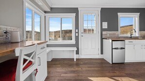 Une cuisine moderne avec des meubles blancs, un plan de travail en bois et de grandes fenêtres offrant une vue sur l’extérieur.