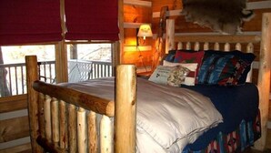 Una habitación de cabaña de madera con una cama, una ventana con cortinas rojas, una lámpara y una cabeza de animal colgada en la pared.