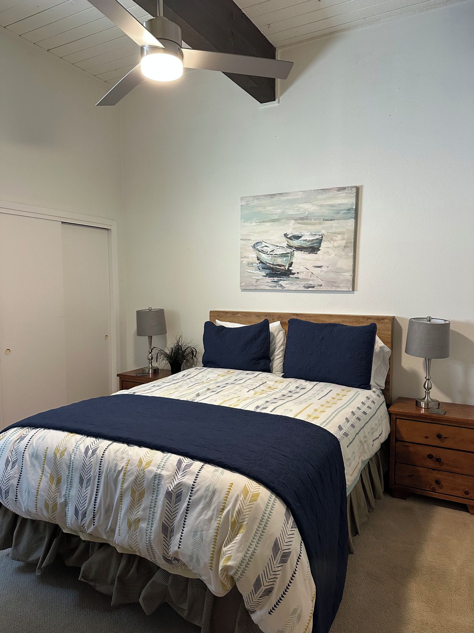Une chambre à coucher avec un lit, deux tables de chevet, un ventilateur de plafond, un tableau et une porte.