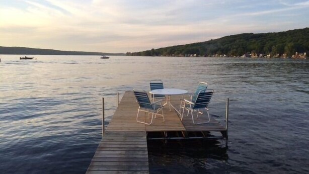 A wooden dock extending into calm water with a table and chairs, surrounded by a natural landscape. - Conesus Lake Cottage Rental (Conesus)