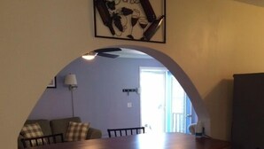 A dining area with a wooden table and two chairs, a sofa with a checkered pillow, and a wall-mounted light fixture. - Conesus Lake Cottage Rental (Conesus)