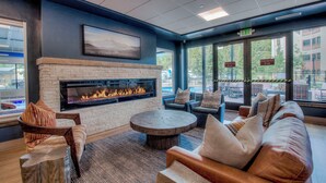 A modern lounge area with a stone fireplace, comfortable seating, and large windows offering views of the outdoors. - Need an escape from summer heat? Cool down in our cozy condo! Summer dates open! (Breckenridge)