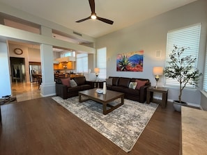 Ein geräumiges Wohnzimmer mit dunklem Holzboden, einem Couchtisch in der Mitte, zwei Sofas mit Kissen, einem Essbereich im Hintergrund und einem Deckenventilator.