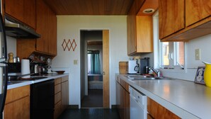 Cocina compacta con armarios de madera, lavavajillas y refrigerador. Un acceso da a un dormitorio con cama y cortinas.