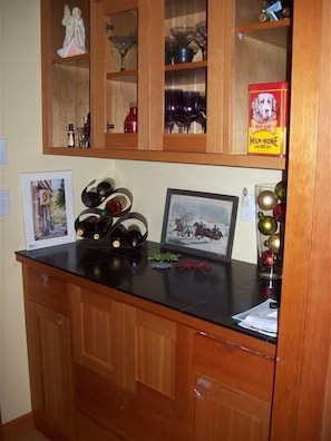 A kitchen with wooden cabinets, a black countertop, and various items like wine bottles, a framed picture, and a box of dog biscuits. - 1600 Sq Ft Craftsman Cottage in the Country (Sequim)