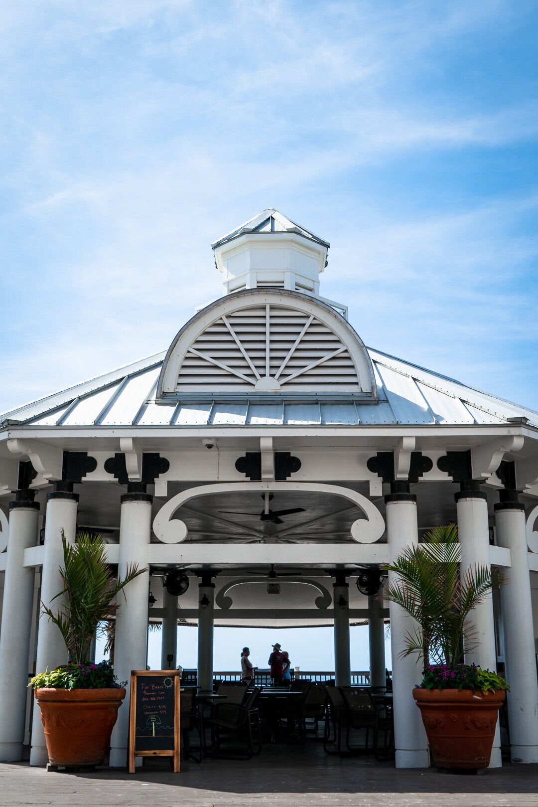 A covered outdoor seating area with a white roof and columns, featuring a signboard and potted plants.