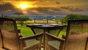 Une table en osier, entourée de chaises, dans un jardin, avec vue sur une plage et un coucher de soleil.