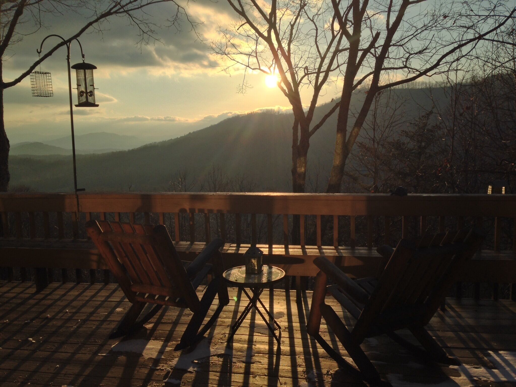 A wooden deck with two chairs and a small table, overlooking a mountainous landscape with a sunset.