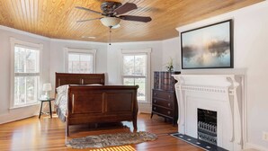 Un dormitorio con techo de madera, una cama, un aparador, una chimenea y ventanas grandes.