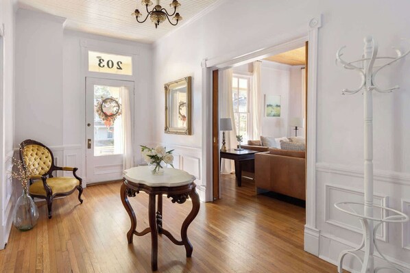 A hallway with a wooden floor, a round table with a vase of flowers, a chair, and a door with a sign.