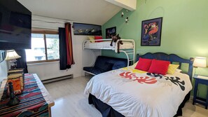 A room with bunk beds, a desk with books, a chair, a bed with colorful bedding, and a window with curtains.