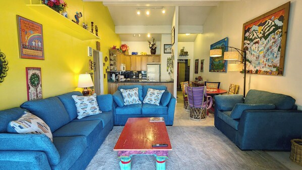 A living room with a blue sofa set, a wooden coffee table, and a kitchen in the background.
