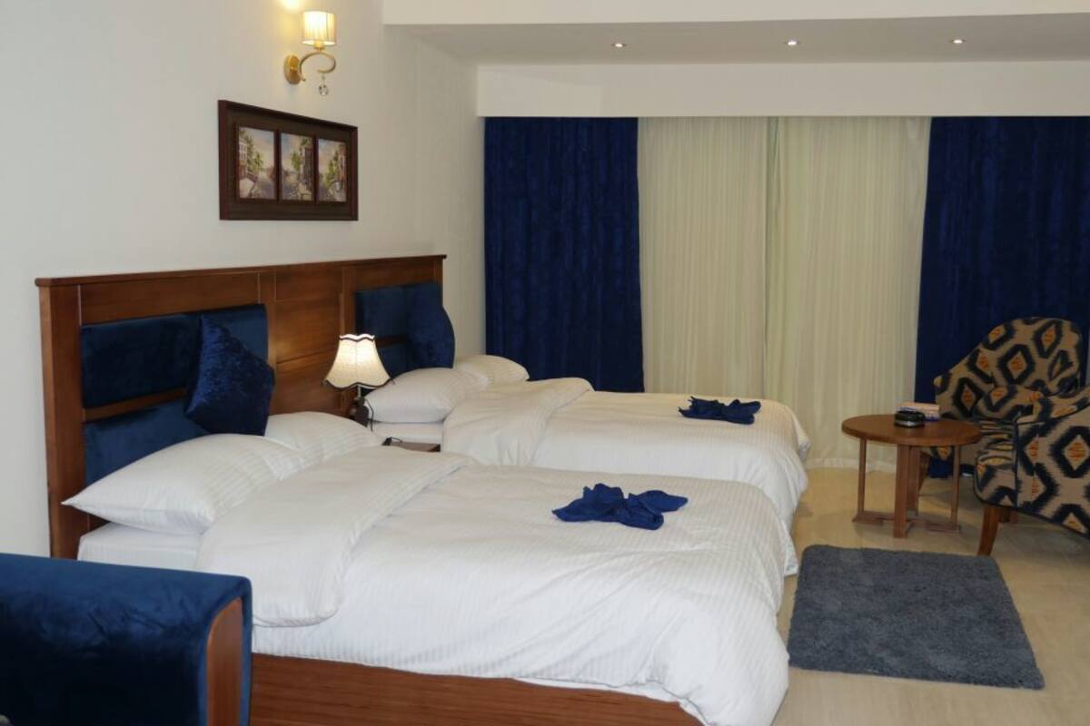 A hotel room with two beds, a small table, a chair, and a view of a building through a window.