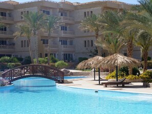A swimming pool with a bridge, palm trees, and a building with balconies in the background.