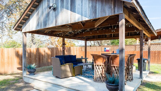 A covered patio with outdoor furniture, a flag, and a wooden fence in the background. - Walk To Main. Hill Country Stone Haus With Unique Outdoor Pavillion (Fredericksburg)