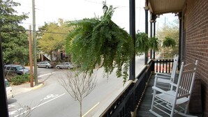 A balcony with a view of a street, a small tree, and a white rocking chair.