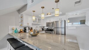 A modern kitchen with white cabinets, a large island with bar stools, stainless steel appliances, and pendant lighting. - Stunning Oceanfront on Crystal Beach with 2 King beds. Beach service included. (Destin)