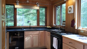 Cucina con mobili in legno, lavastoviglie nera, piano cottura e lavello. Ampie finestre offrono vista sull'esterno.