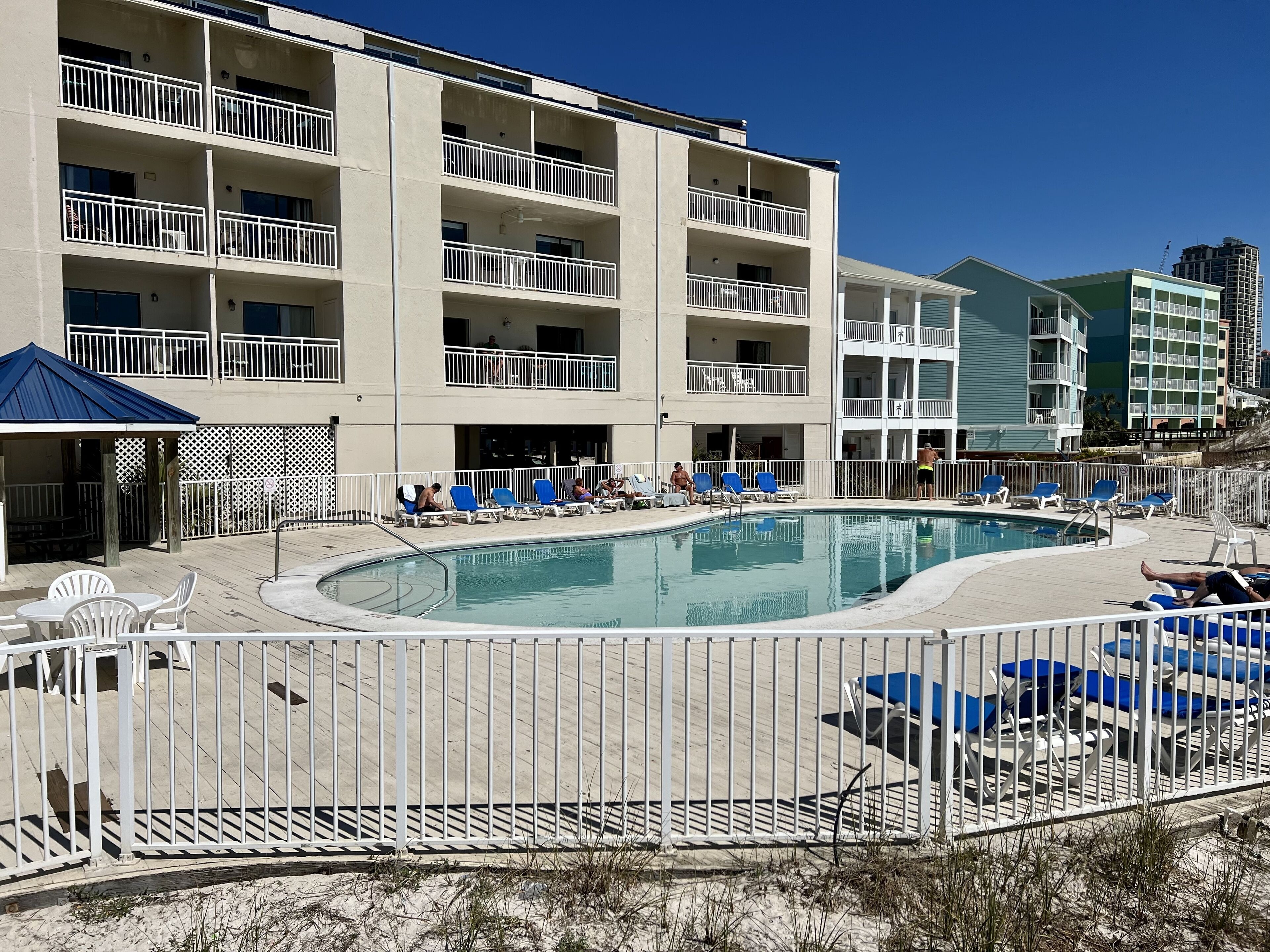 A multi-story hotel with a swimming pool surrounded by blue lounge chairs and a fenced deck area.
